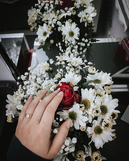 A close-up of a floral bouquet featuring white daisies with yellow centers, accented by delicate sprigs of baby's breath, and a single red rose nestled among the daisies. The bouquet is held by a pers