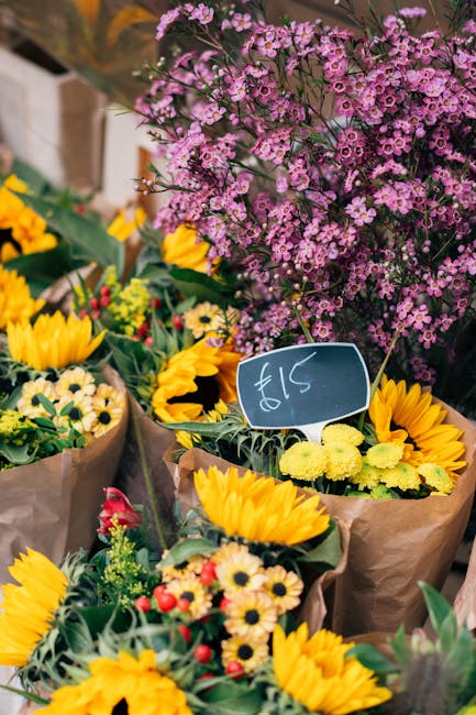 A vibrant outdoor display of fresh floral arrangements featuring bright yellow sunflowers, white daisies with yellow centers, and small red and pink blossoms, all wrapped in brown paper and set agains