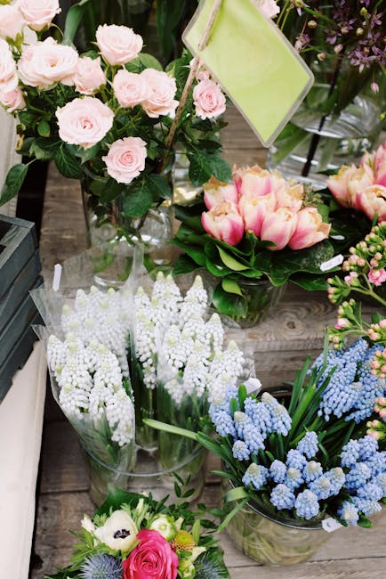 A display of fresh flowers on a wooden surface, featuring a bouquet of light pink roses with lush green foliage in a clear glass vase, alongside pink and white tulips arranged in a separate transparen