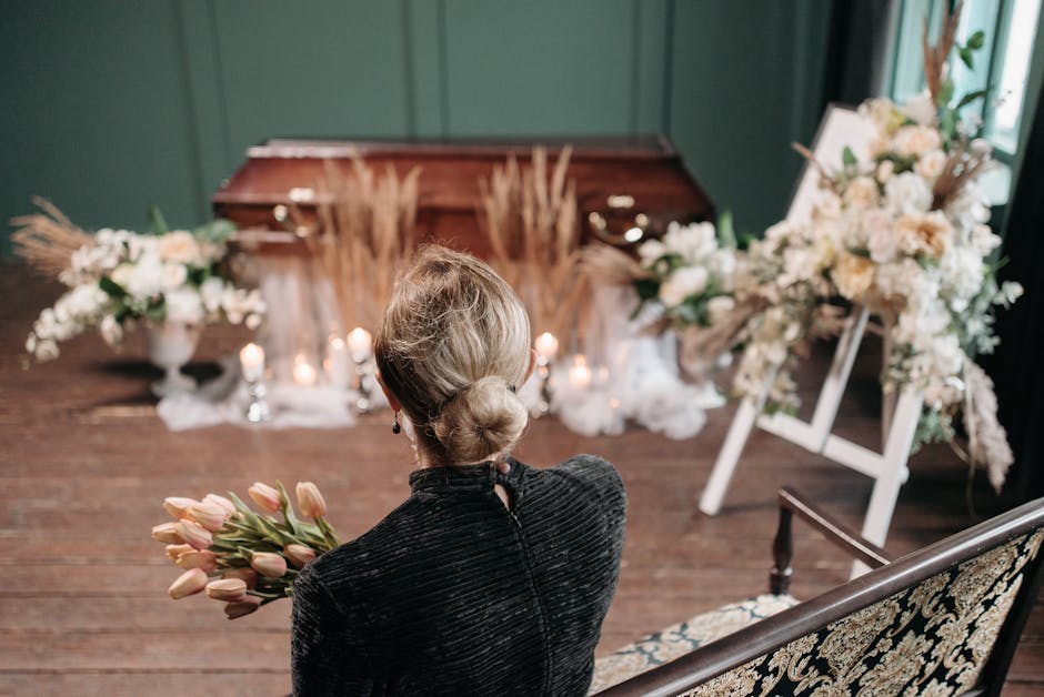 A woman with blonde hair styled in a low bun, wearing a black textured dress, holds a bouquet of pink tulips, standing in front of a floral arrangement for a funeral. The background features a wooden 