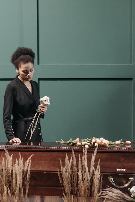 A young woman with dark curly hair, dressed in a black coat, holds a small white flower with a pink center in her left hand while standing next to a wooden table. The table displays a flower arrangeme
