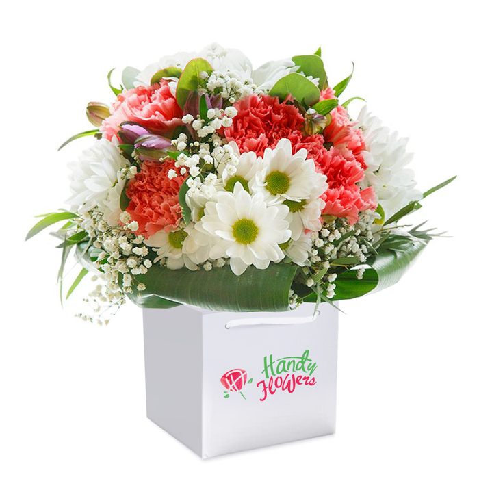 With striking clarity, this bouquet invites admiration from within its square, snowy-white gift bag-ready to delight Leytonstone residents from Bushwood to Hollow Ponds. At its core are lush coral-pink and bold red carnations, recognizable by their full, deeply frilled petals, creating a sumptuous palette and tactile richness. Pops of crisp white daisy-like blossoms with vivid lime-green centers brighten the scene, while the addition of frothy white baby's breath intermingles airily among the larger blooms, lending a whimsical delicacy. Nestled discreetly at the side, unopened purple-streaked buds contribute intrigue and subtle colour. The cluster is embraced by abundant, glossy dark green leaves, which form a supportive coupe and natural highlight to the vivid blooms. The bag's elegant ribbon handles glisten, suggesting the bouquet's easy portability across Leytonstone's bustling streets or tranquil green corners. The overall impression is one of freshness and craftsmanship-a florist's celebration of both beauty and local spirit, perfect for gifting at any special occasion where the thoughtful artistry of Leytonstone shines.
