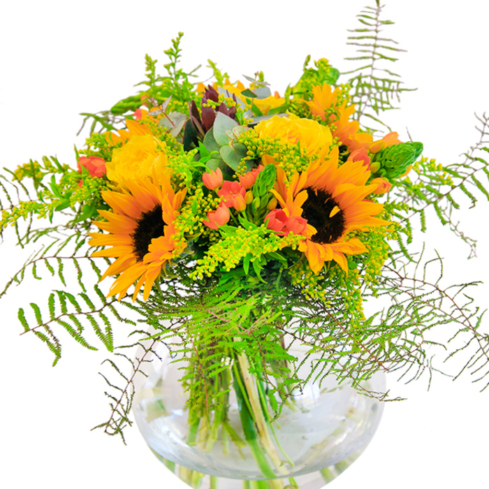 This sun-kissed bouquet, a staple of Leytonstone's artisan floristry, glows with a joyful duet of sunflowers and golden yellow roses artfully arranged in a round, crystal-clear glass vase. The arrangement feels like a nostalgic nod to summers in Wanstead Park: bold sunflowers, their rich brown centres and expansive yellow petals, command attention with a vibrant, open-hearted energy. Woven beside them, tightly coiled yellow garden roses bloom in plush rosettes, their silky petals adding elegance and softness. Red Hypericum berries, like scattered jewels, glisten amidst the greenery, lending unexpected charm and vivid colour pops that echo the local community's creative flair. Lush ferns and airy, green filler soften the bouquet's bold lines; slender goldenrod and gentle eucalyptus bring variety and movement, contributing subtle hues of green and dusty blue. The vase's clarity showcases every fresh stem and water's gentle ripple, heightening the sense of natural beauty and simplicity found in Leytonstone's favourite gathering spots. An ideal centrepiece for milestone events, client meetings, or simply delivering a sunlit welcome to a neighbour, the bouquet's scent mixes the whisper of cut greens with the warming sweetness of garden blooms. Each detail, from textural contrasts to the gentle play of light and shadow, celebrates Leytonstone's blend of tradition and modern spirit.